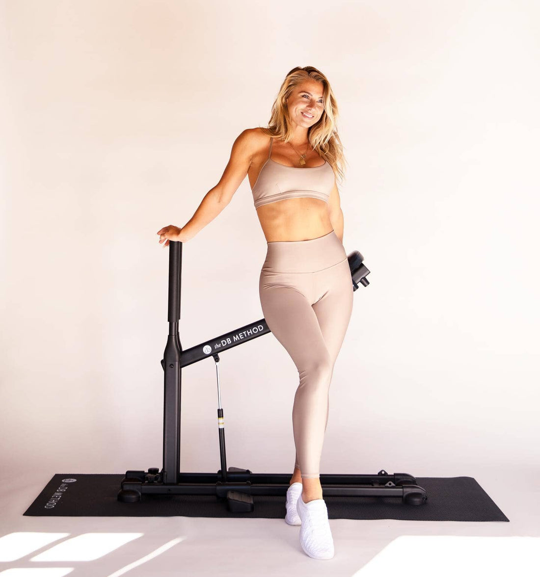 A smiling woman stands beside The DB Method Squat Machine on a workout mat.