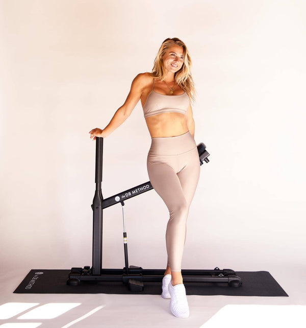 A smiling woman stands beside The DB Method Squat Machine on a workout mat.