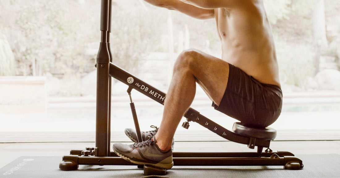  Shirtless man performing a seated squat on The DB Method machine in a bright home setting.