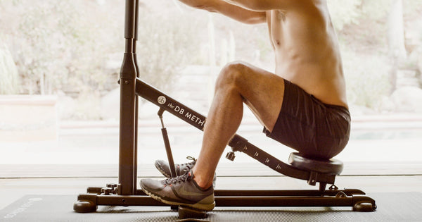  Shirtless man performing a seated squat on The DB Method machine in a bright home setting.