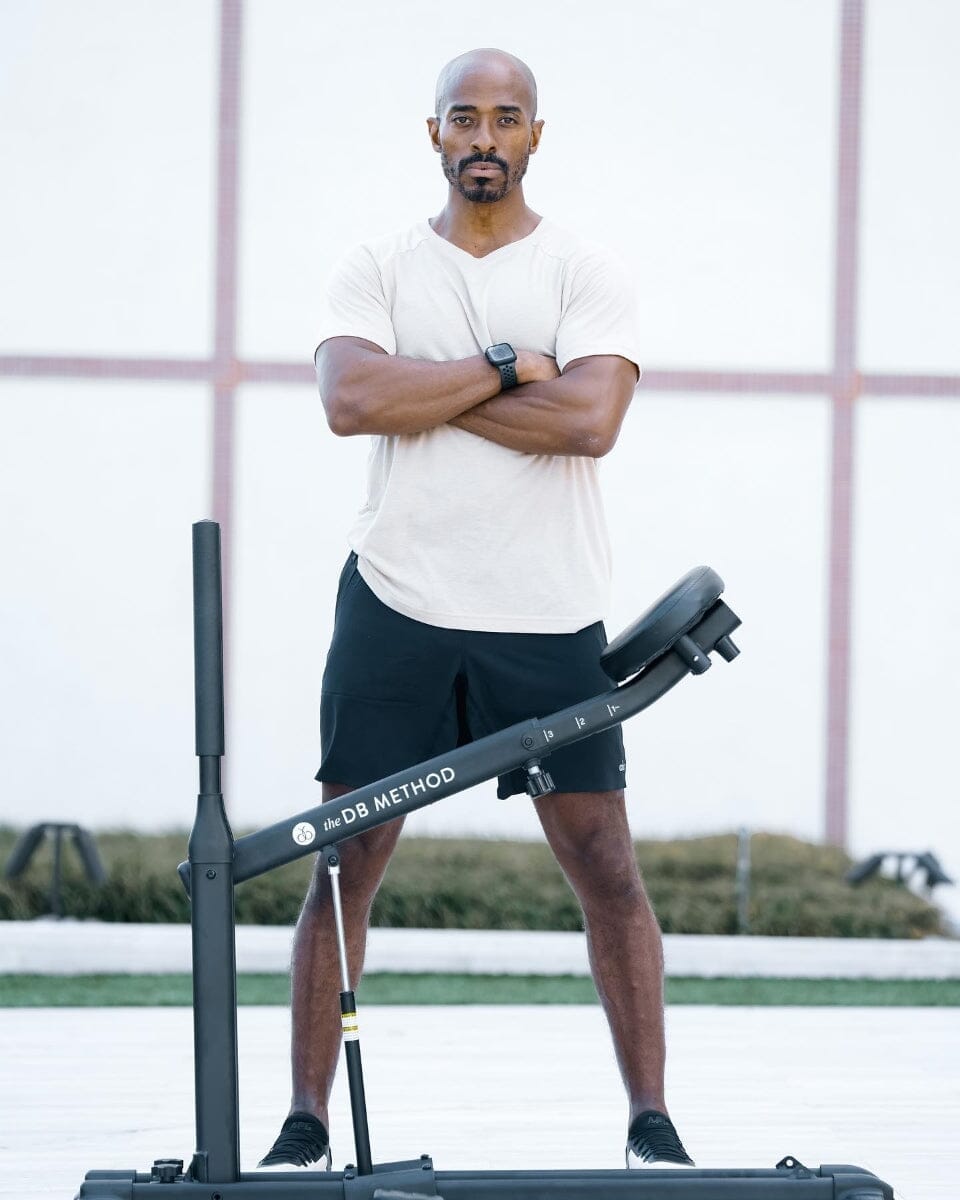 A fit man stands with arms crossed behind The DB Method machine, wearing a light tee and black shorts.