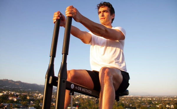 Man using The DB Method squat machine outdoors against a skyline, with the brand logo visible.