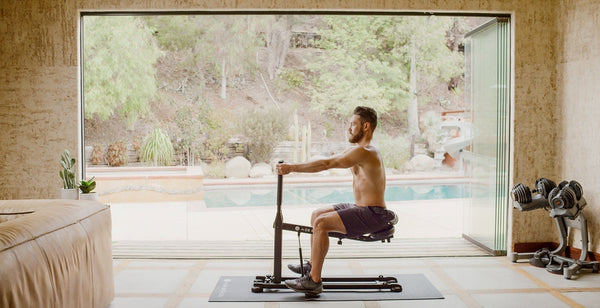 A man uses The DB Method Squat Machine on a workout mat in a bright home gym.