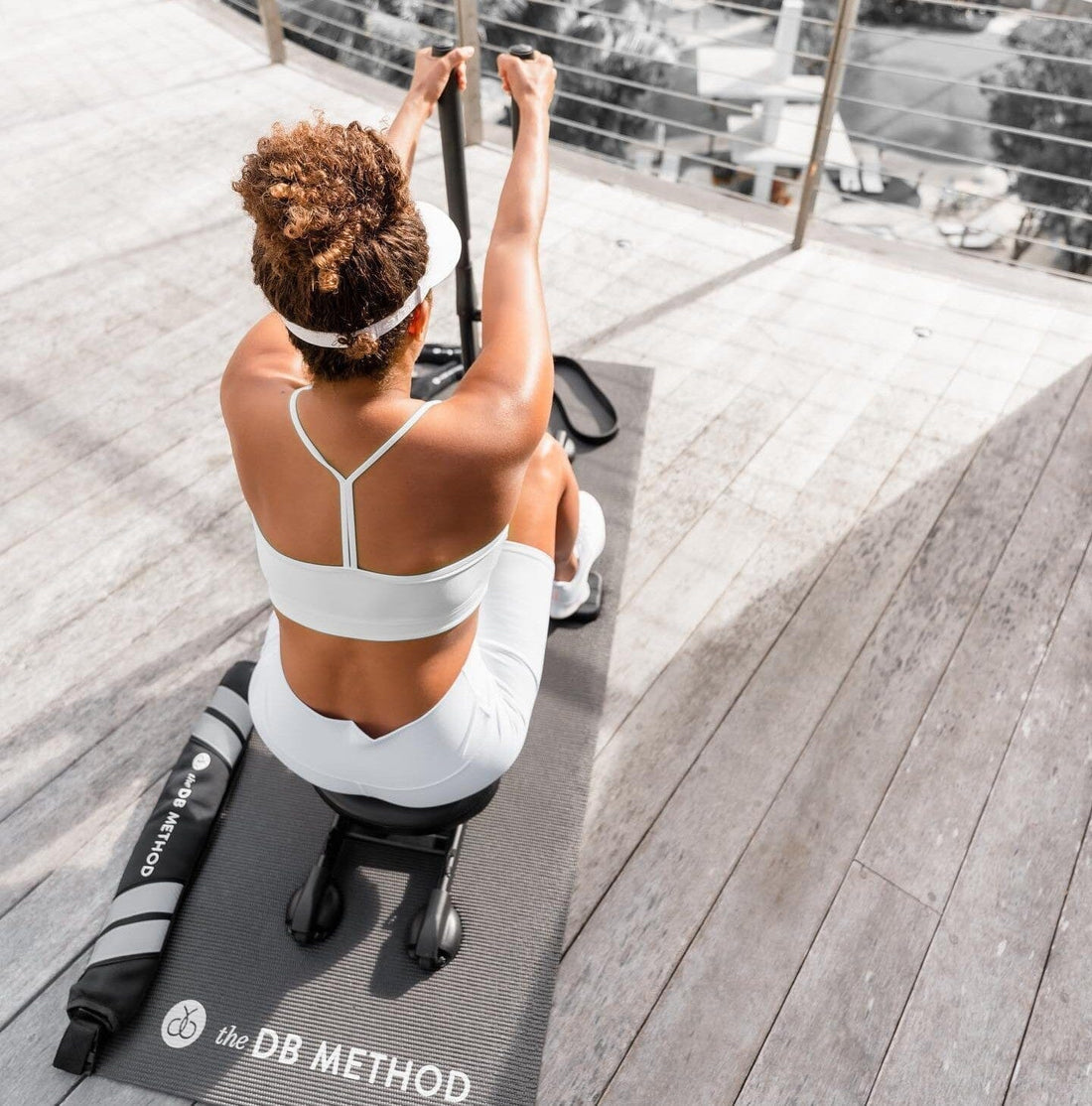 Woman working out outdoors using The DB Method machine, seated on branded mat in bright light.