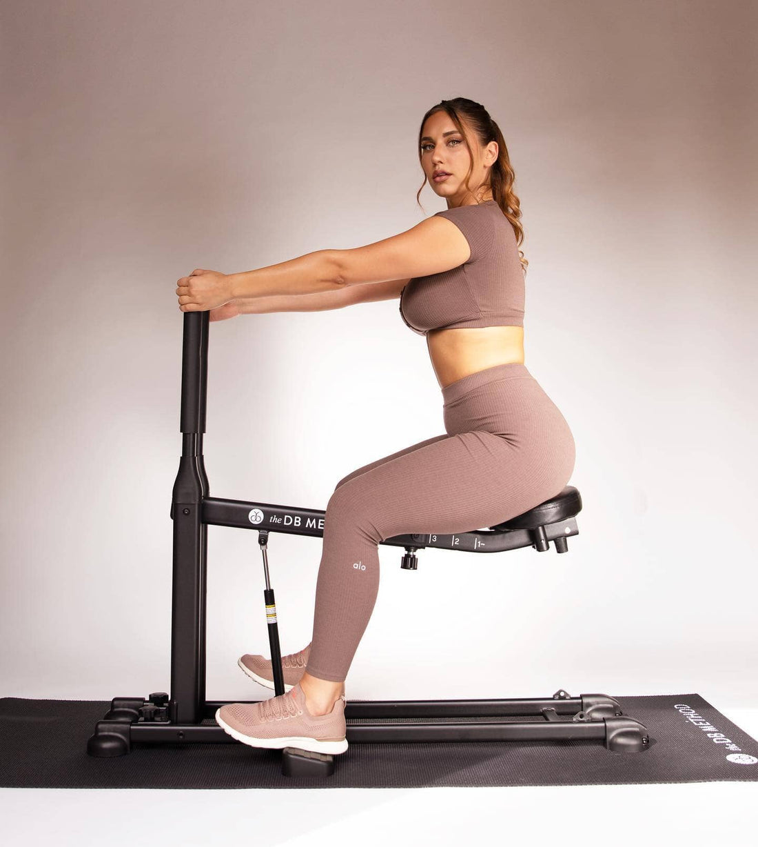 Woman seated on The DB Method machine, wearing a matching athletic set and sneakers, holding the front handlebar.