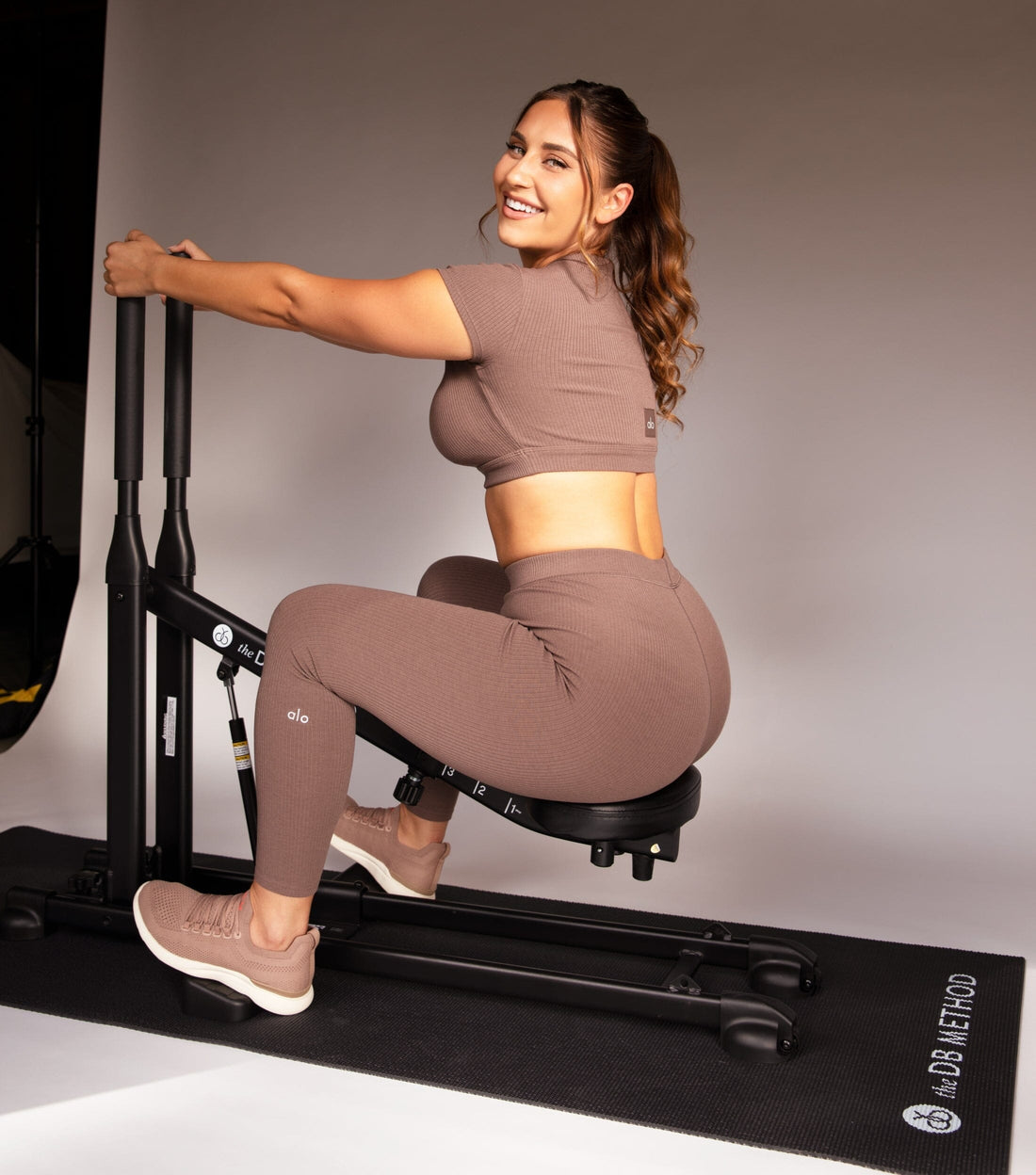 Woman seated on The DB Method squat machine in a studio, wearing a brown athletic set and sneakers.