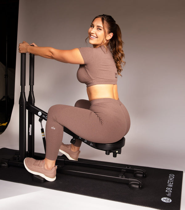Woman seated on The DB Method squat machine in a studio, wearing a brown athletic set and sneakers.