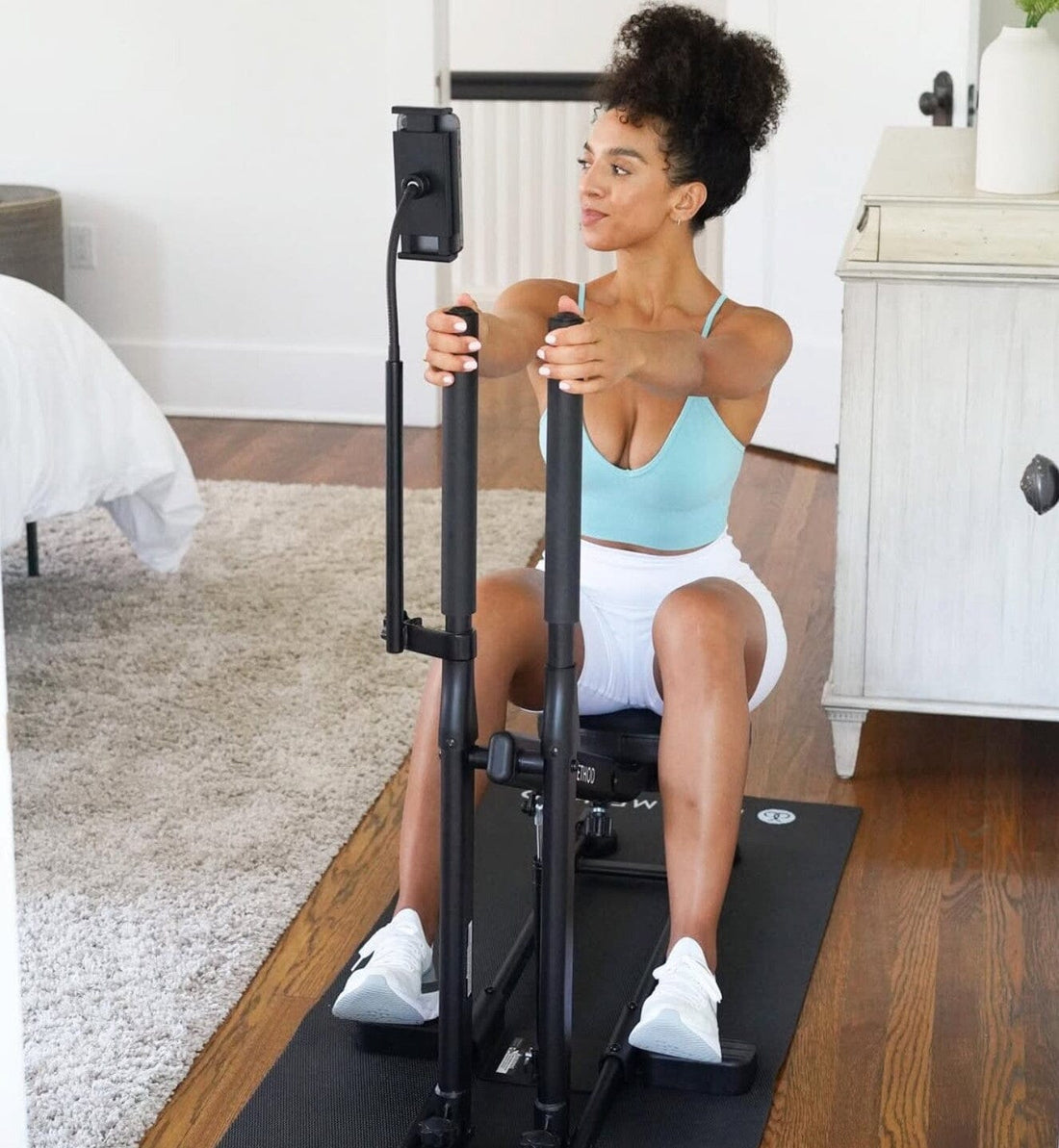 Woman sitting on The DB Method machine indoors, wearing a light blue sports top, white shorts, and sneakers.