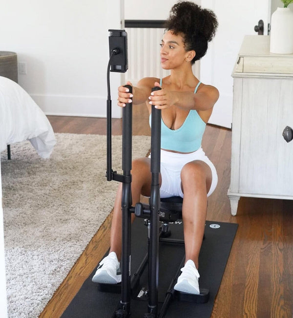 Woman sitting on The DB Method machine indoors, wearing a light blue sports top, white shorts, and sneakers.
